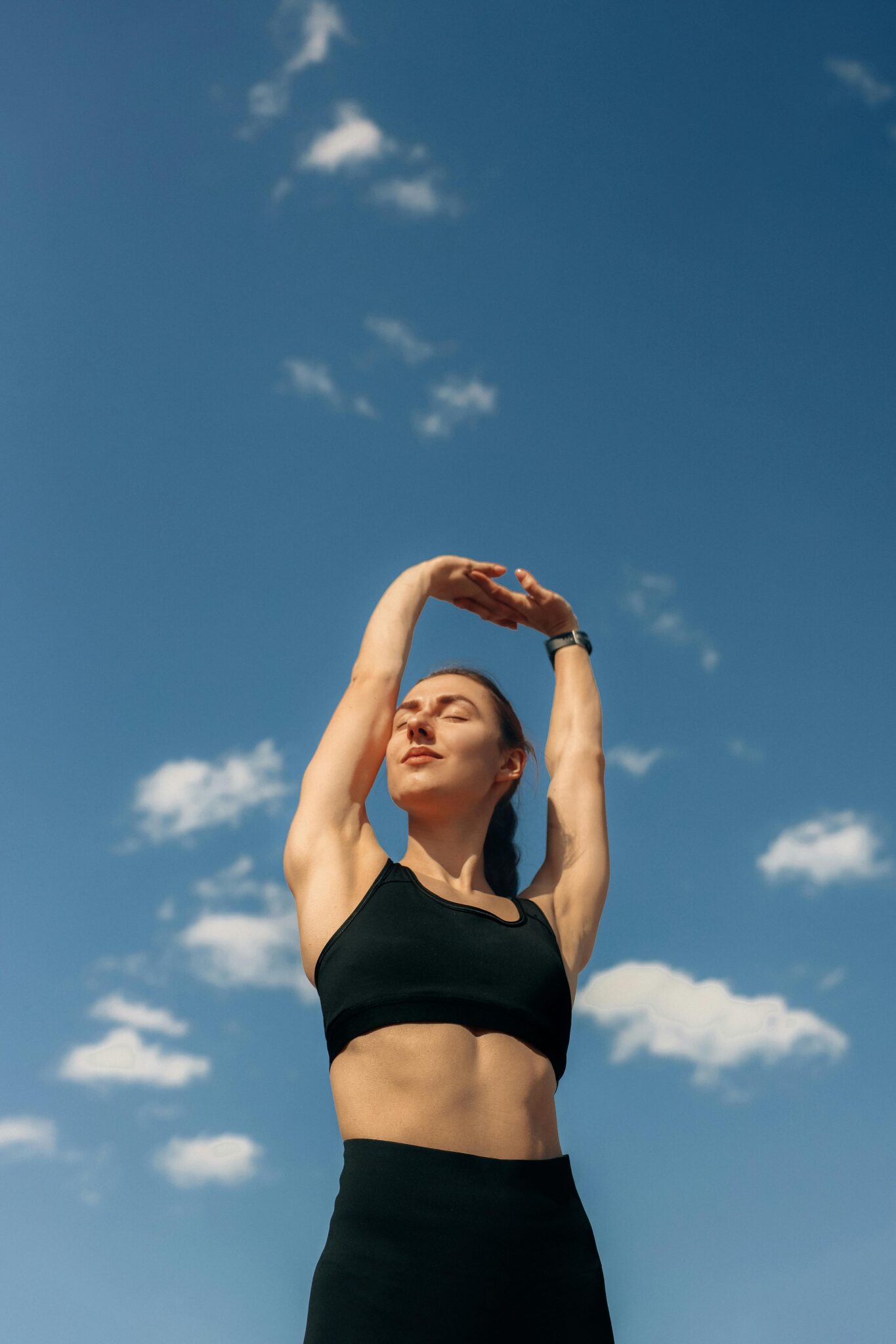 active health sport and A woman in black leggings and sports bra stretches under a clear blue sky, promoting fitness and freedom.