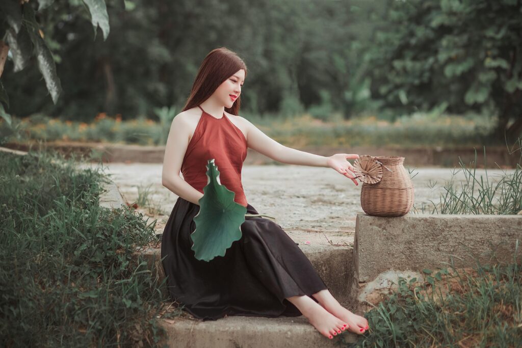 Serene young woman sitting outdoors holding a lotus leaf next to a wicker basket with barefoot.