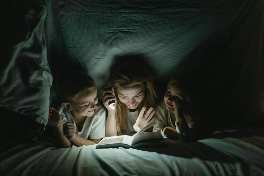 A mother shares a perfect bedtime routine story with her kids under a cozy blanket, lit by a flashlight.