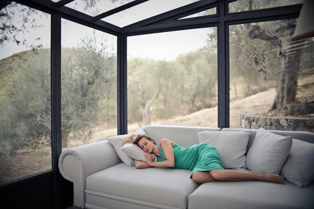 A woman in green dress power naps on a sofa in a glass-walled room with olive trees outside.