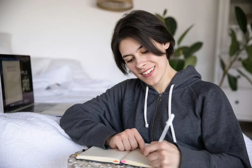 A woman in a hoodie sits on bed writing journaling in a notebook, capturing a candid moment.