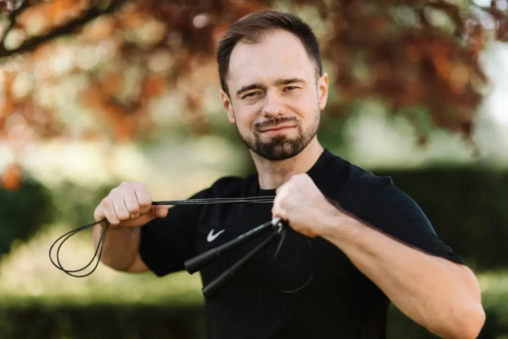 Bearded man in a black shirt confidently posing with a jump rope outdoors.