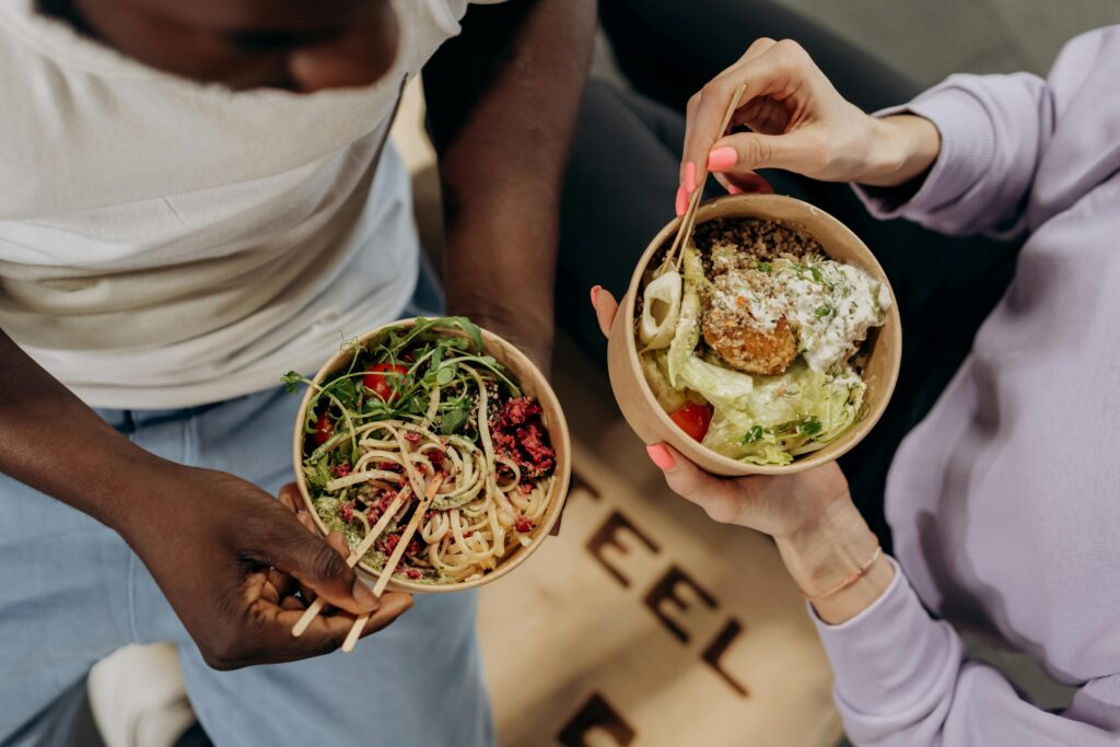 Two people holding timing proteins bowls filled with salad and pasta, emphasizing healthy lifestyle choices.