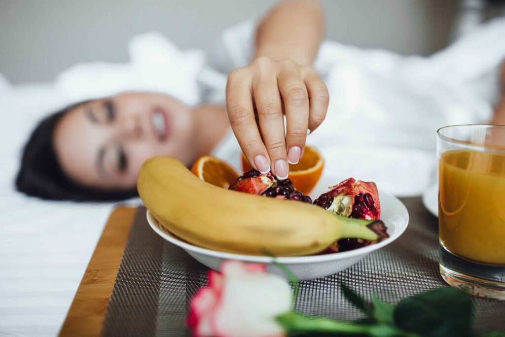 Cropped beautiful brunette woman lying in morning bed, tray with orange juice, banana, pineapple, apples, soft morning warmth
